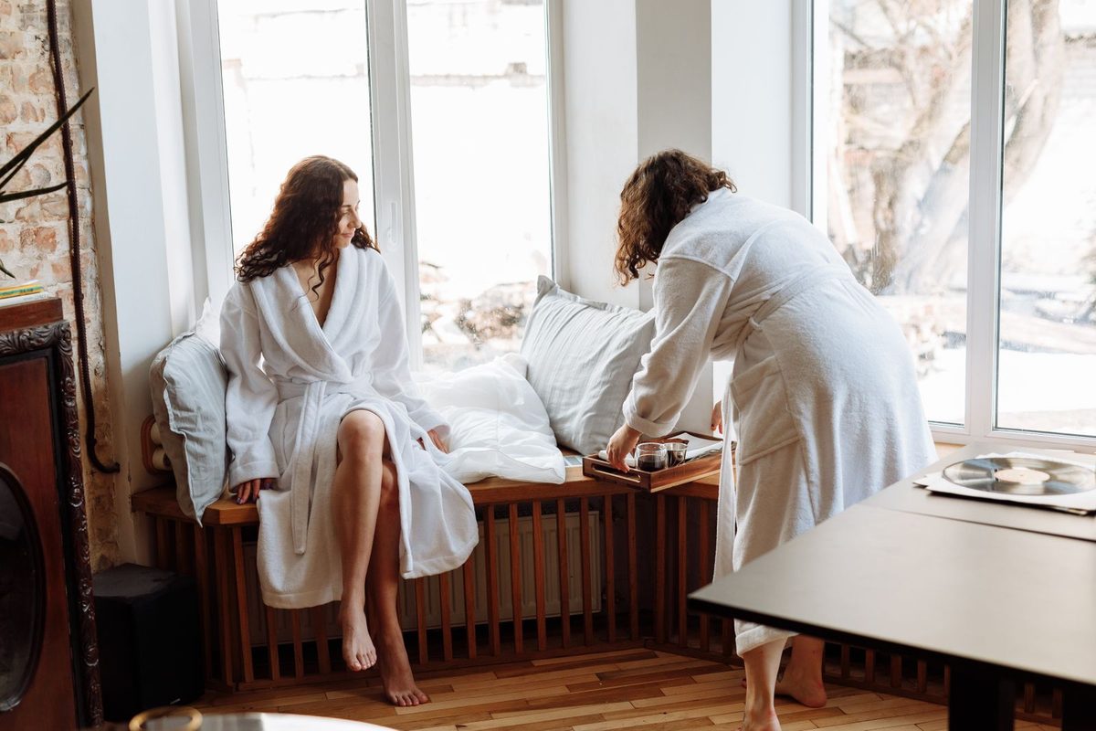 Two people in white robes at a sunny window seat with coffee